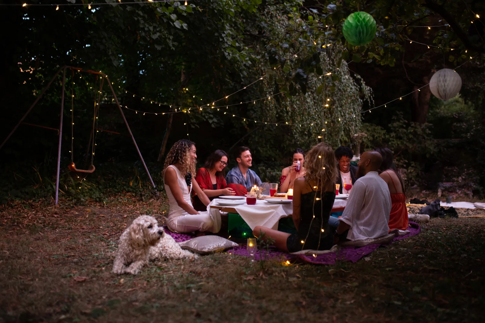 Friends gathered around a candlelit table in a London park at dusk, with string lights overhead and a dog resting on the rug