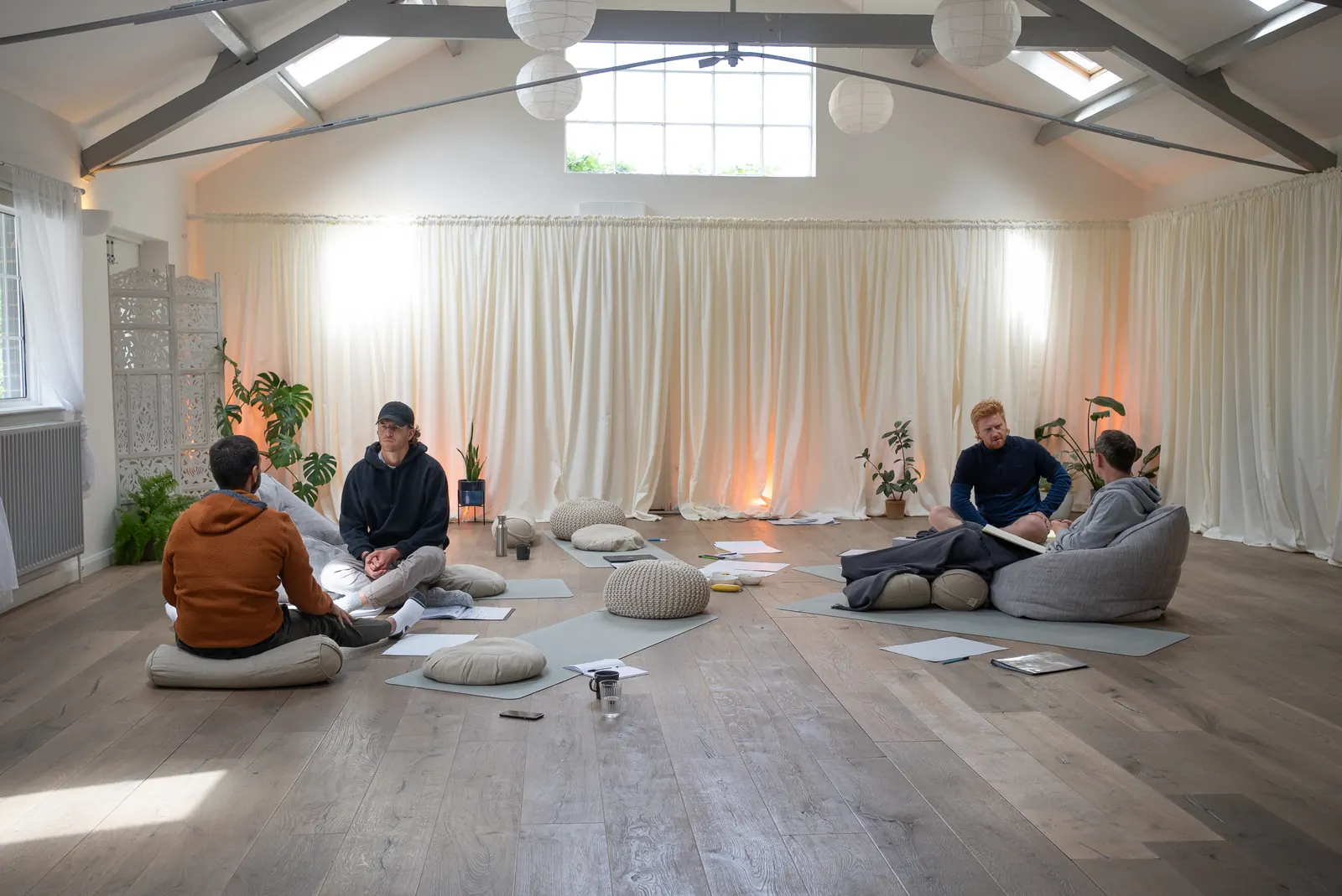 Wide view of a full training circle, five men seated on cushions facing each other on the floor of a studio